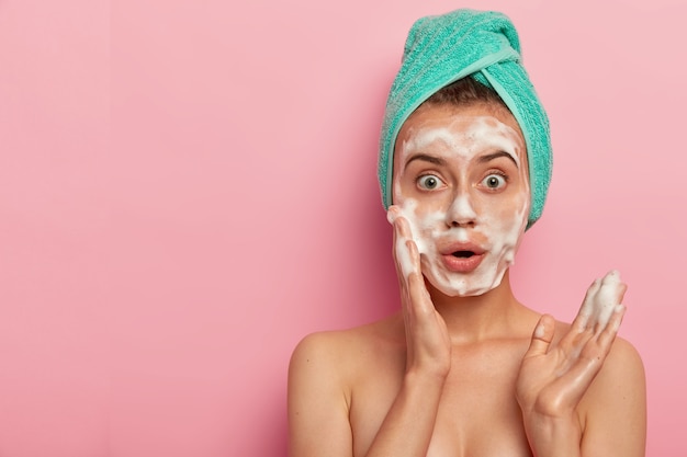 Photo of surprised European woman washes face with foam gel, wants to have refreshed well cared skin, stands topless, wears wrapped towel on wet hair, poses against pink background, free space aside
