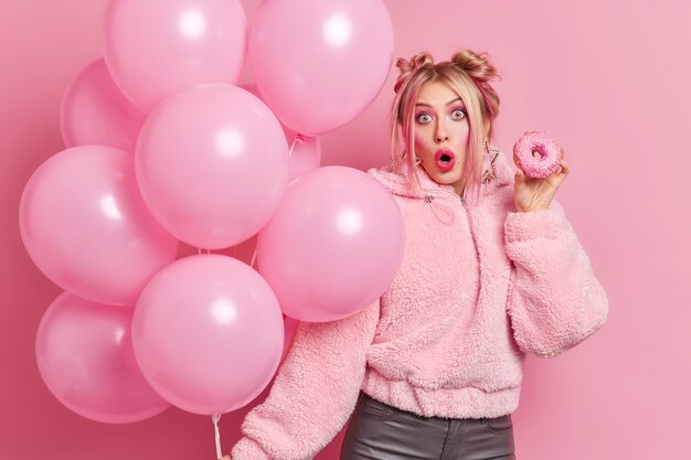 Photo of surprised European woman with hairstyle wears fashionable clothes holds delicious doughnut and balloons enjoys birthday party shocked to get unexpected gift models indoor. Everything in pink