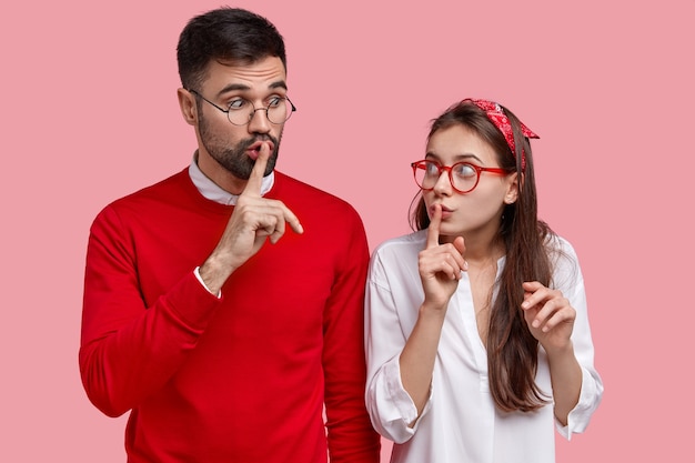 Free photo photo of surprised female and male gossip together, make silence gesture, tell secret information, look at each other with conspiracy