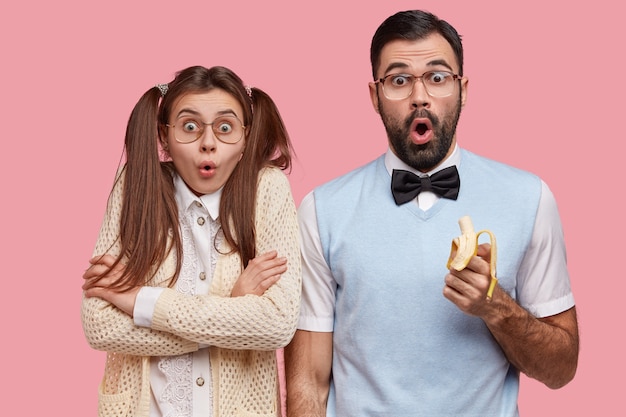 Free photo photo of surprised female and male wonks stare with disbelief, eat delicious banana, dressed in old fashionable clothes