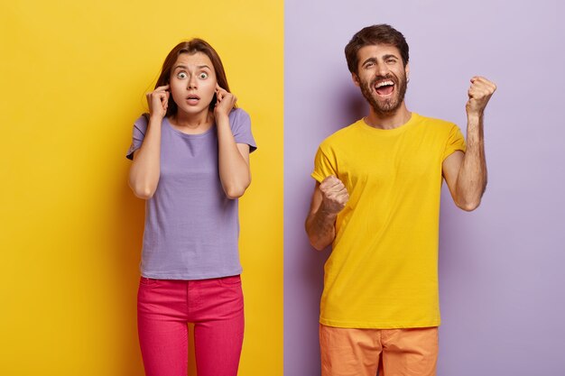 Photo of surprised female plugs ears, shocked with noise, glad man clenches fists