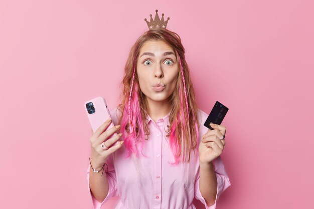 Photo of surprised long haired woman keeps lips folded wears crown and shirt makes payment online via smartphone application holds banking card poses against pink background buys things in store