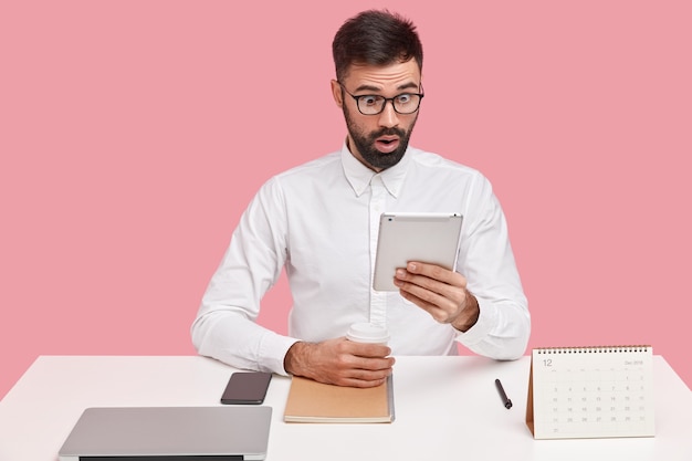 На фото удивленный сотрудник-мужчина смотрит на экран тачпада, удивляется, читает шокирующее сообщение, пьет кофе из одноразовой чашки, носит очки