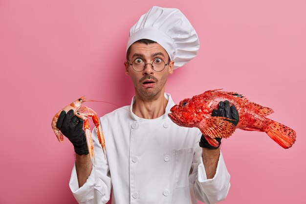 Photo of surprised man cook holds crefish and red sea bass, going to prepare dish from seafood, tries best recipe, stands at kitchen, dressed in white uniform