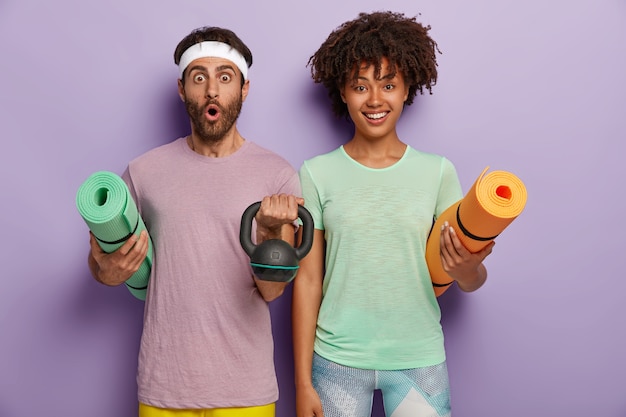 La foto dell'uomo sorpreso tiene la stuoia e il peso, indossa la fascia e la maglietta, la donna dalla pelle scura allegra trasporta il tappetino fitness, pronta per l'allenamento con l'allenatore