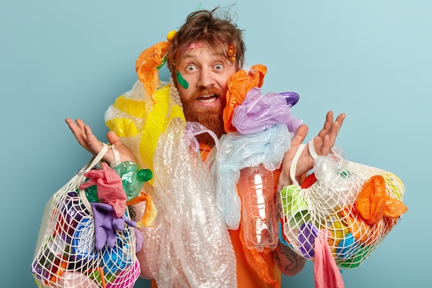 На фото удивленный рыжеволосый мужчина с густой бородой, перегружен мусором, собирает пластик