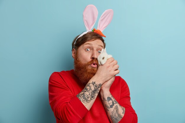 Photo of surprised redhead man keeps small animal near face, plays with white rabbit, wears bunny ears, prepares for costume party on Easter eve, poses against blue wall. Spring holiday