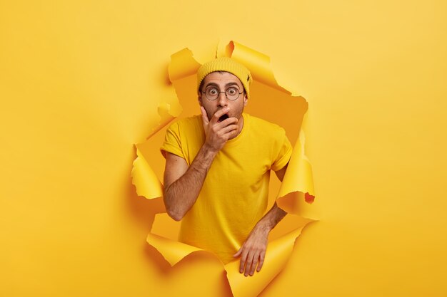 Photo of surprised scared man breaks through color paper wall, covers mouth, has stupefied expression