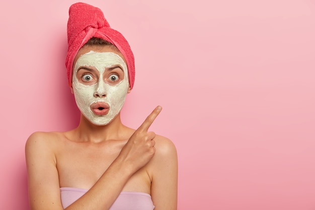 Фотография удивленной безмолвной женщины в маске для лица, у которой косметические процедуры дома, шокированное выражение лица, показывает указательным пальцем в сторону, влажные волосы обернуты полотенцем.