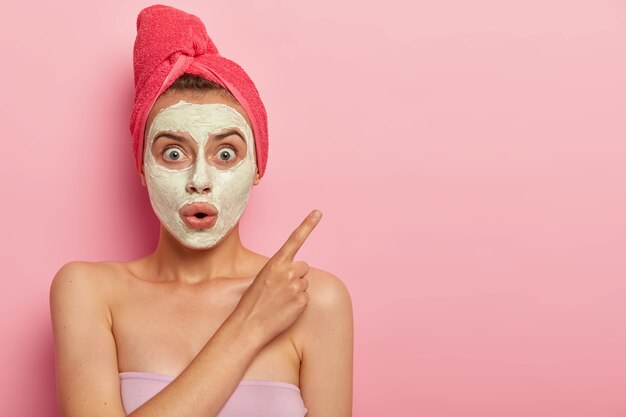 Photo of surprised speechless woman wears facial mask, has beauty procedures at home, shocked expression, points aside with index finger, wrapped towel on wet hair