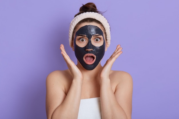 Photo of surprised speechless woman with opened mouth, wears mud facial mask, has beauty procedures, girl with shocked expression, wrapped towel on body, isolated on lilac wall, keeps palms near face.