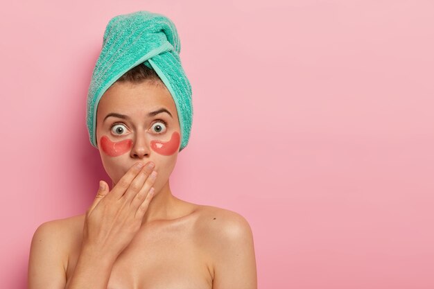 Photo of surprised woman has omg expression, covers mouth with palm, cares about complexion, applies collagen patches for reducing dark circles under eyes