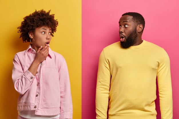 Photo of surprised woman and man look with shock at each other, feel embarrassed after hearing bad news, cannot believe their eyes, dressed casually, isolated over pink and yellow wall