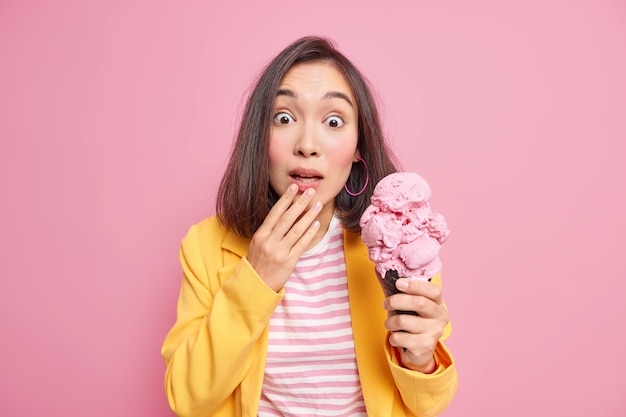 Photo of surprised young Asian woman with dark hair stares shocked dressed in striped t shirt and yellow jacket holds yummy ice cream isolated over pink wall. People and emotions concept