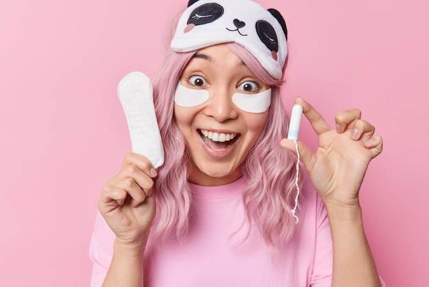 Photo of surprised young Asian woman with toothy smile looks happily holds tampon and sanitary napkin chooses better hygiene product wears sleepmask after sleeping isolated over pink background