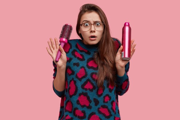 Photo of surprised young European woman with puzzled expression, wears big spectacles and casual sweater, holds comb with hairspray