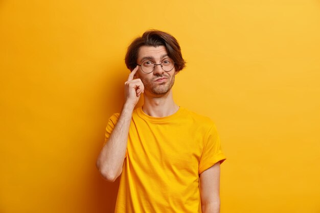 Photo of thoughful young European keeps finger on temple imagines something wears round spectacles and casual t shirt isolated over yellow  wall makes important decision picks variant
