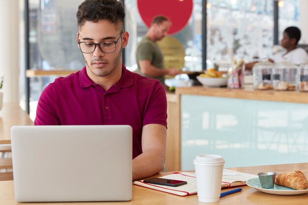 Photo of thoughtful bearded man works on creative ideas for publication, keyboards information on portable laptop computer, spends time in coffee shop, has delicious snack with croissant and tea