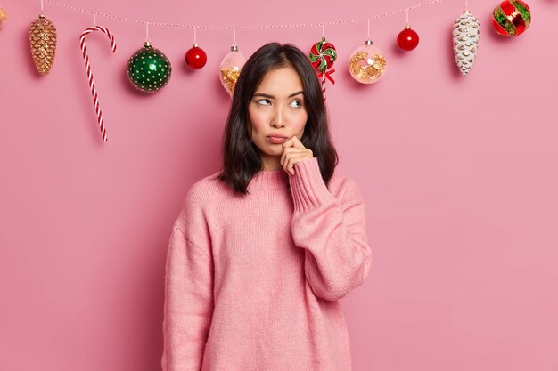 Фото задумчивой брюнетки азиатской женщины стоит недовольно одетая в повседневный джемпер держит руку на лице думает об украшении дома перед новогодними позами