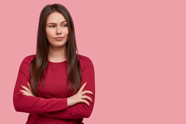 Photo of thoughtful brunette young woman with dark hair, keeps arms folded, considers something in mind, wears red sweater, stands against pink background. People