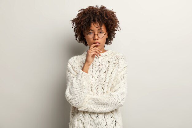 Photo of thoughtful dark skinned female has curly hair, serious look, being deep in thoughts, looks straightly, wears long sleeve sweater, isolated over white wall. Thinking concept