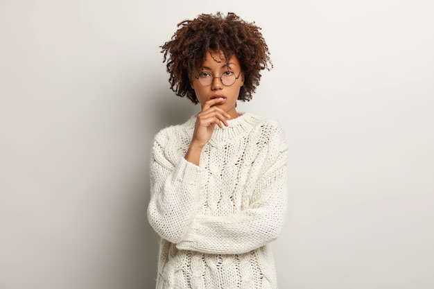 На фото задумчивая темнокожая женщина, вьющиеся волосы, серьезный вид, задумчивая, прямолинейно, в свитере с длинным рукавом, изолирована на белой стене. Концепция мышления