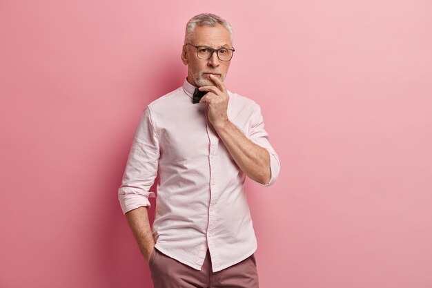 Photo of thoughtful elderly man keeps hand on mouth, other hand in pocket of trousers, has pensive expression