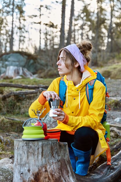 Photo of thoughtful female has coffee at scenic spot, poses near stamp with portable camp stove and coffee maker