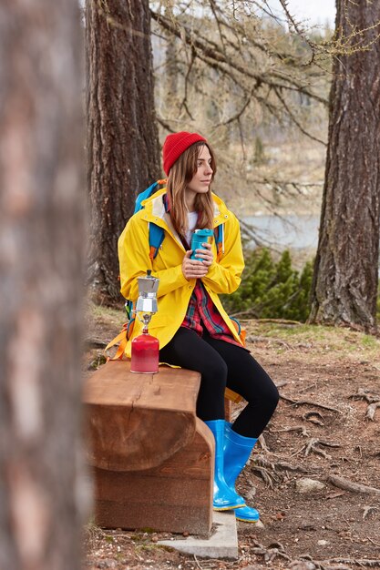 Фотография вдумчивой путешественницы отдыхает на деревянной скамейке в лесу, пьет чай из термоса, варит кофе на походной плите, носит красную шляпу