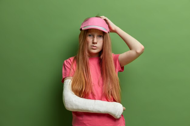 Photo of thoughtful indecisive woman scratches head and tries to remember all details about accident happened with her, has broken arm in gypsum, dressed casually, isolated on green wall