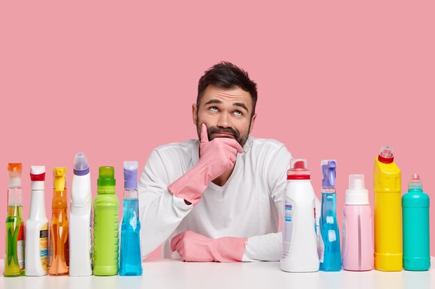 Photo of thoughtful man holds chin, looks pensively upwards, wears white jumper and gloves, uses washing up liquid, cleanser, isolated over pink space