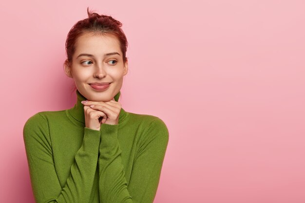 photo of thoughtful pleasant looking european girl keeps both hands under chin, looks away, ressed in green casual jumper, isolated on pink wall, free space aside