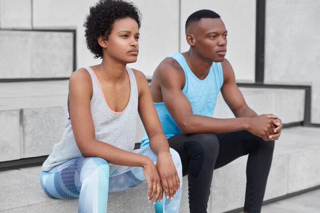 Photo of thoughtful sporty dark skinned woman and man rests after exercises on stairs, have motivation for staying healthy and fit, dressed in active wear, have workout together. Athletic trainings