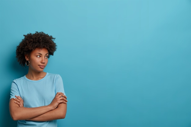 Photo of thoughtful woman has healthy skin, keeps hands crossed over chest, concentrated aside, wears casual t shirt, isolated on blue wall, blank space for your information, makes plans