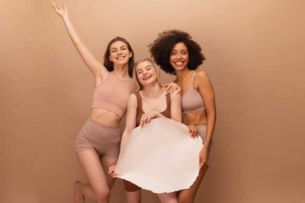 ベージュの背景に空白の白いプラカードを保持している気分の良い3人の幸せな若い異人種間の女性の写真