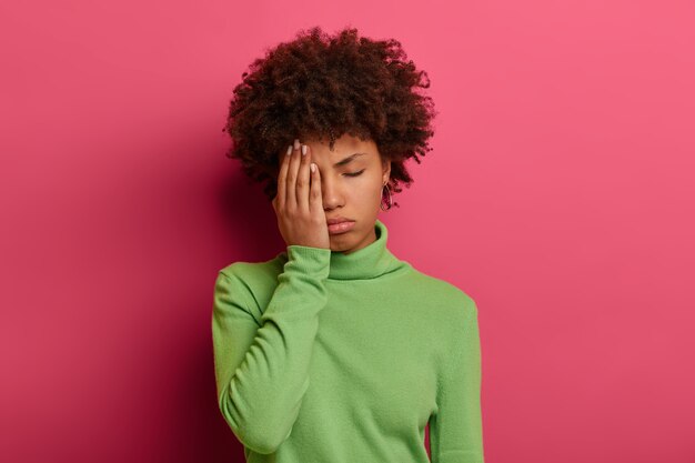 Photo of tired Afro American woman covers half of face with palm, keeps eyes closed, wants to sleep, needs rest