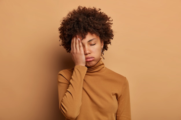 Free photo photo of tired curly woman covers face with palm, feels overworked and fatigue, wants to sleep, tilts head, wears casual turtleneck