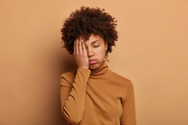 Photo of tired curly woman covers face with palm, feels overworked and fatigue, wants to sleep, tilts head, wears casual turtleneck