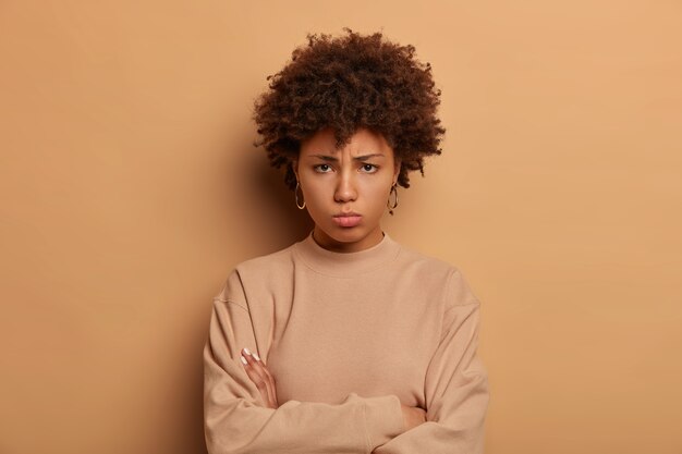 Photo of troubled sad woman stands upset and offended, keeps arms folded, looks with regret from under forehead, wears casual beige jumper, expresses negative emotions, blows cheeks from anger