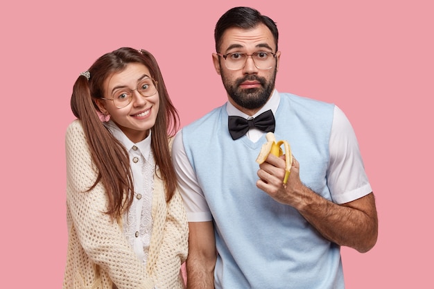 Photo of two clever cheerful friends meet together during day off, pose at camera, eat banana, wear spectacles