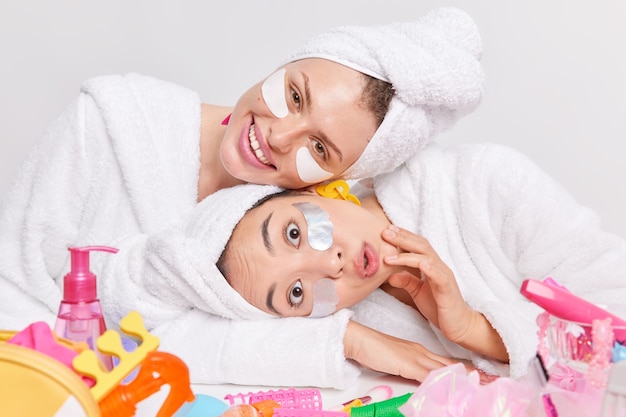 Photo of two mixed race young women tilt heads pose near table with cosmetic products apply patches under eyes