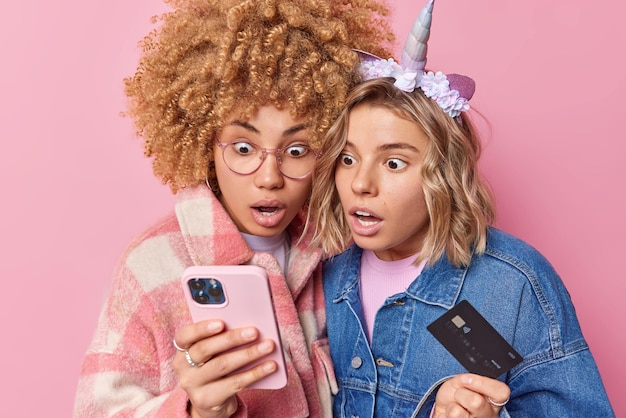 Фотография двух потрясенных красивых женщин, смотрящих на смартфон, держащих кредитную карту, не ожидающих получить бонус и кэшбэк, сделать онлайн-платеж, одетых в модную одежду, изолированных на розовом фоне
