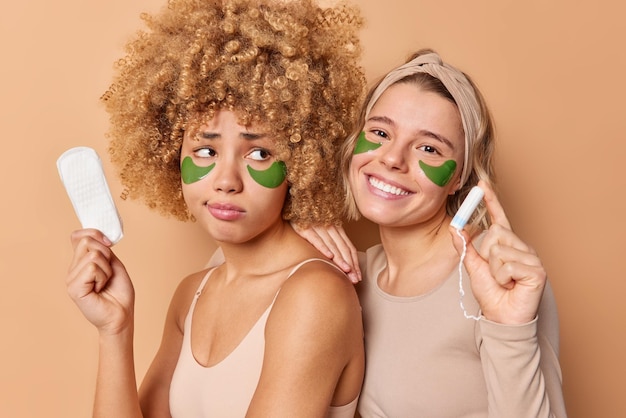Free photo photo of two women friends pose with hygiene products choose between sanitary napkin and tampon apply hydrogel patches under eyes undergo beauty procedures stand closely to each other indoors