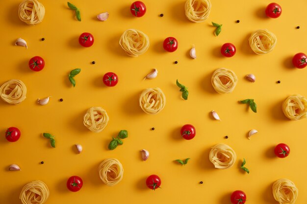 Photo of uncooked pasta nests lying around edible red tomatoes, garlic, peppercorns, basil on yellow background. Cooking nourishing meal. Italian traditional cuisine. Big variety of products