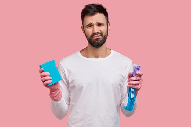 Photo of unhappy Caucasian man with dark stubble, dressed in white clothes, holds mop and washing spray, prepares for spring cleaning