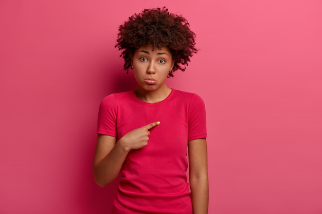 Photo of unhappy young African American woman points at herself with miserable expression, asks why am I guilty, purses lower lip from bad emotions, wears crimson t shirt, sad being mentioned