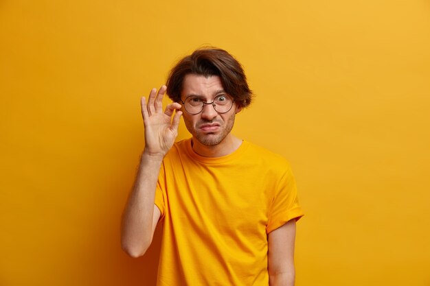 Photo of unimpressed displeased adult man shapes very tiny object, demonstrates something little, has scrupulous look , wears round transparent glasses, isolated on yellow wall