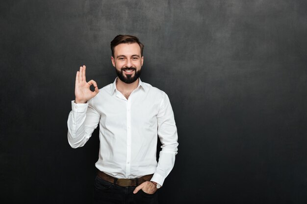 Photo of unshaved guy in office smiling and gesturing with OK sign expressing everything is alright, isolated over graphite