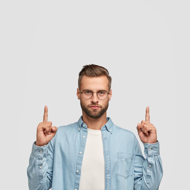 Photo of unshaven hipster dressed in casual shirt, points with both index fingers upwards.