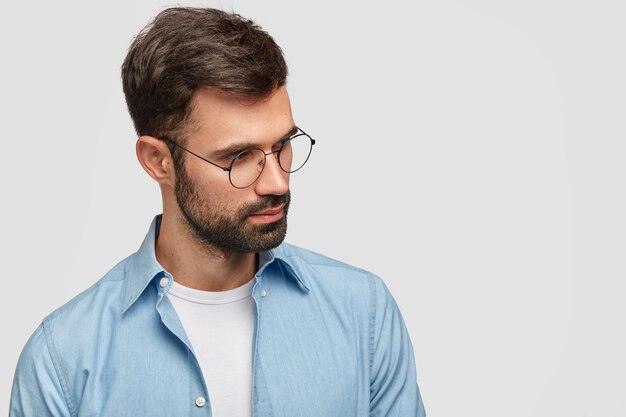 Photo of unshaven young male with dark stubble and hair, wears spectacles and shirt, focused aside, poses against white wall